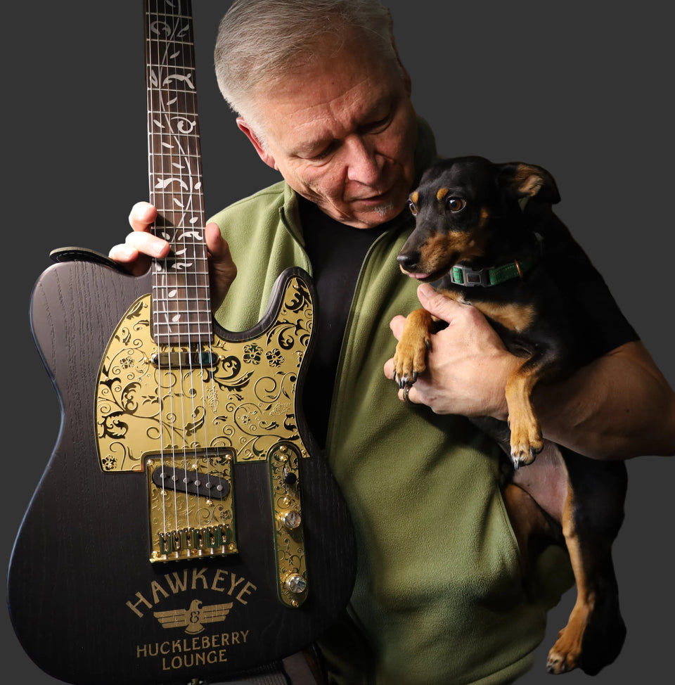 Man holding a guitar and a small dog in a production room.
